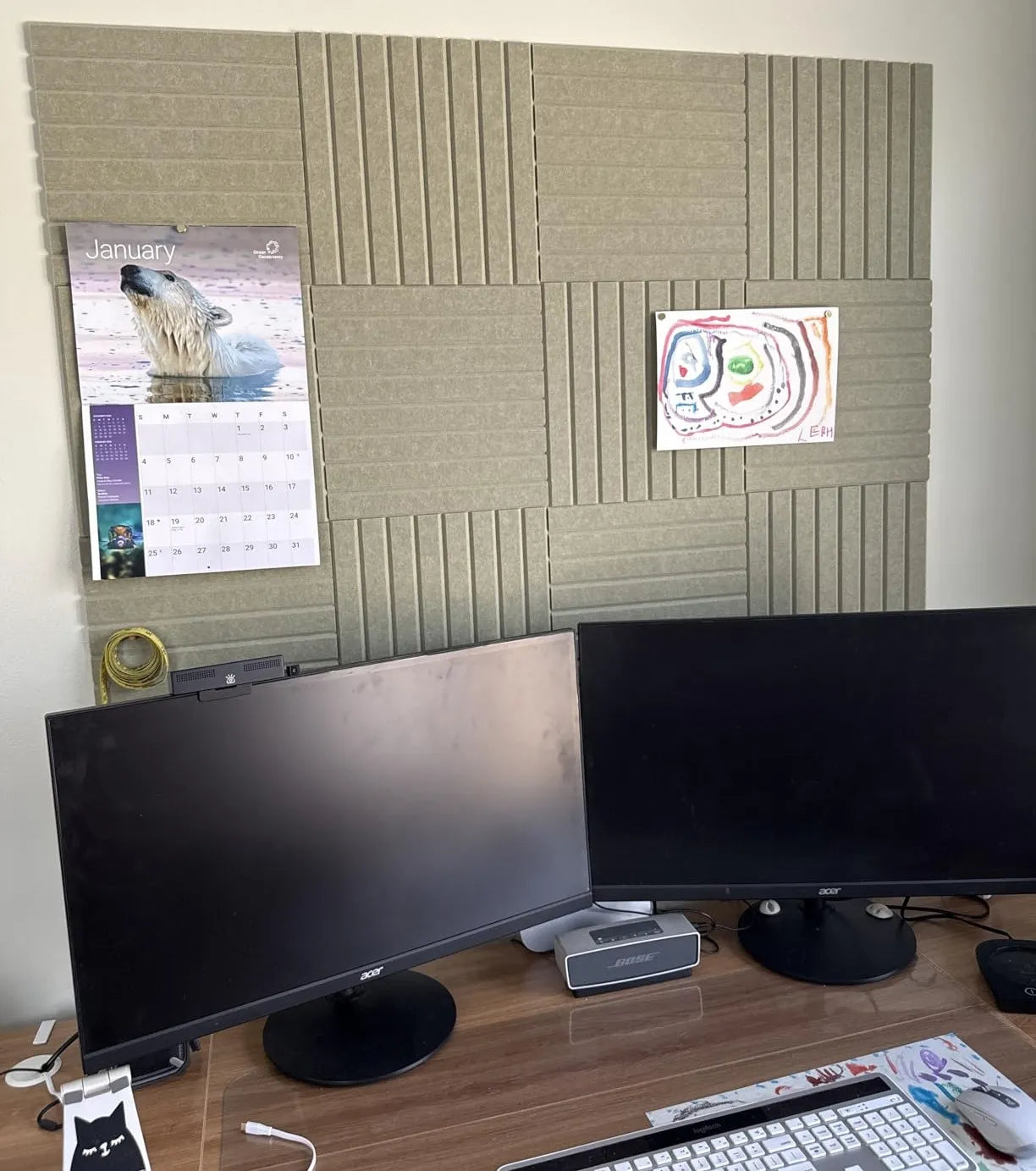 Large felt bulletin board wall covered in sage tiles used as pin board behind dual monitor desk setup